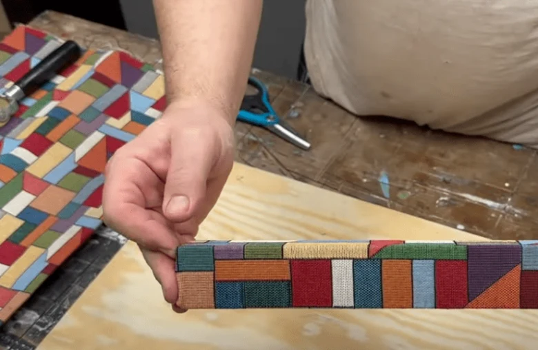 Ruka drží rámeček nábytku obložený barevnou látkou s geometrickým vzorem.