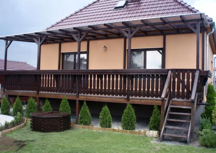 Dřevěný balkon u domu s schodištěm a keři.