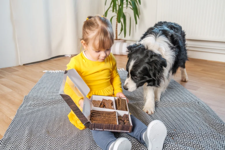 Dívka s psem sedí na koberci, otevřená krabice psích sušenek.