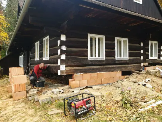 Muž opravuje základy staré roubené chalupy s cihlami.