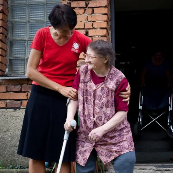 Mladá osoba pomáhá seniorce s holí ven z budovy.