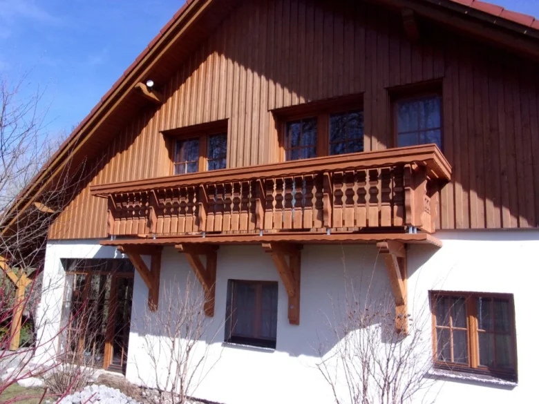 Dřevěný balkon na fasádě domu s bílou omítkou.